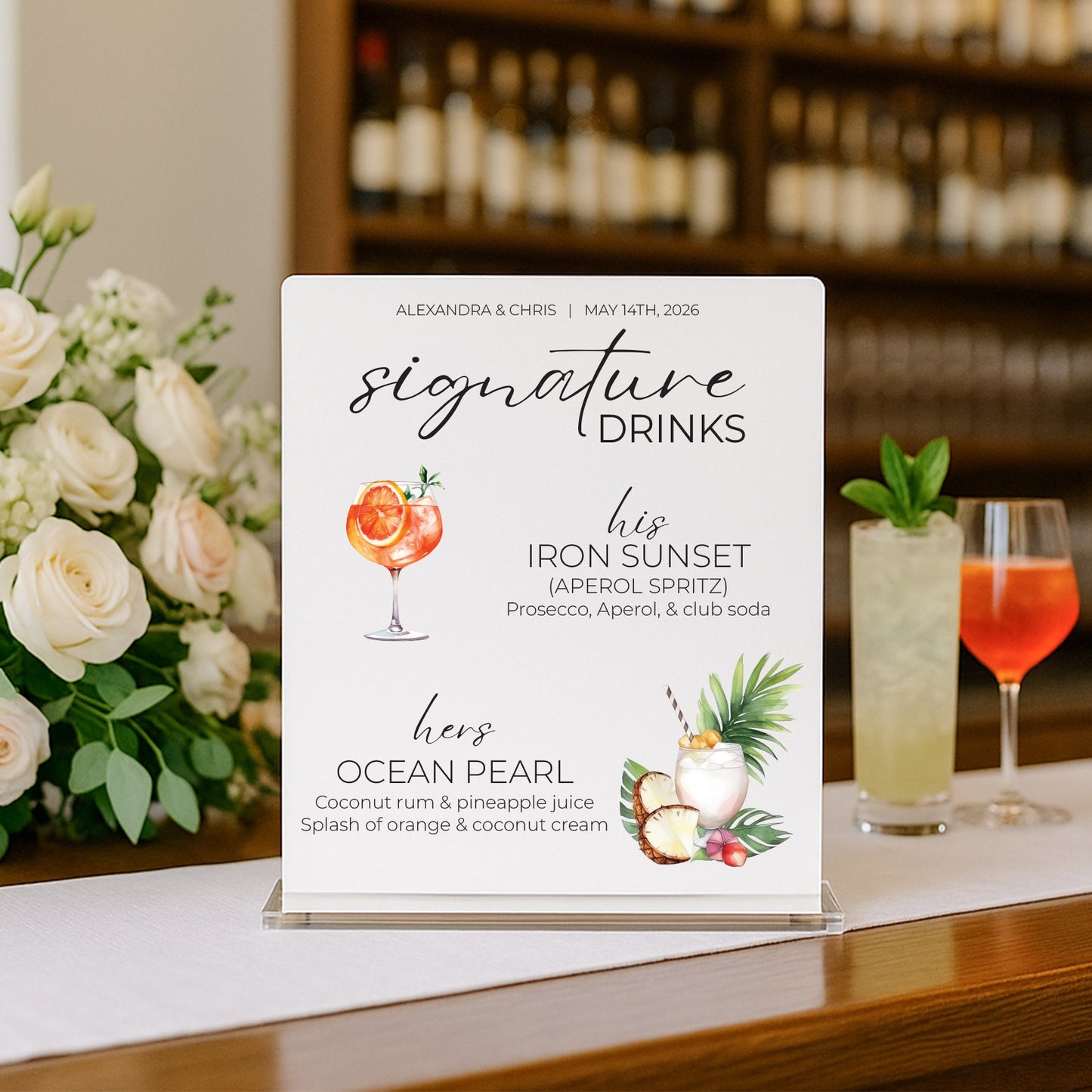 A wedding cocktail menu sign displayed on a bar counter, featuring elegant black script text, floral illustrations, and two prepared cocktails in the background with a blurred bar shelf.