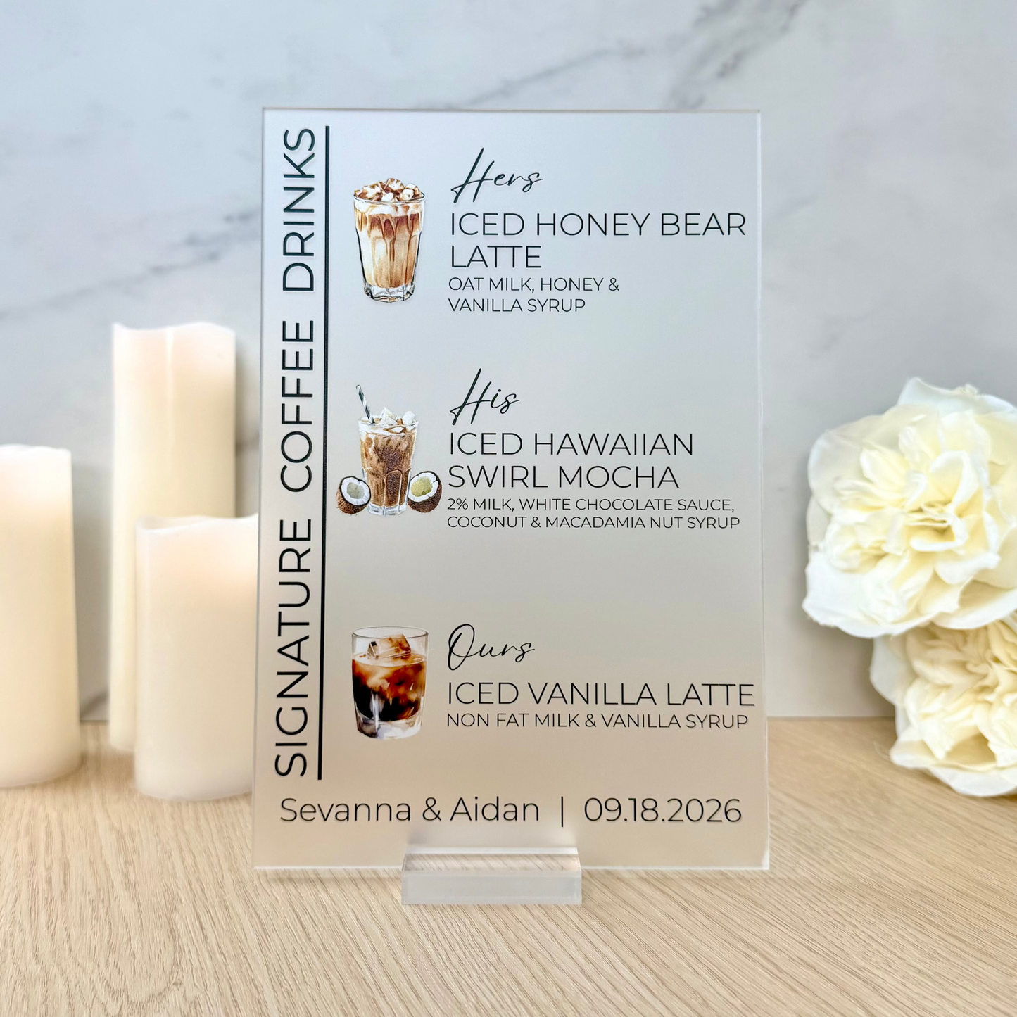 A clear acrylic custom coffee drink menu card standing on a wooden table, flanked by three white pillar candles on the left and white roses on the right, against a light marble wall.