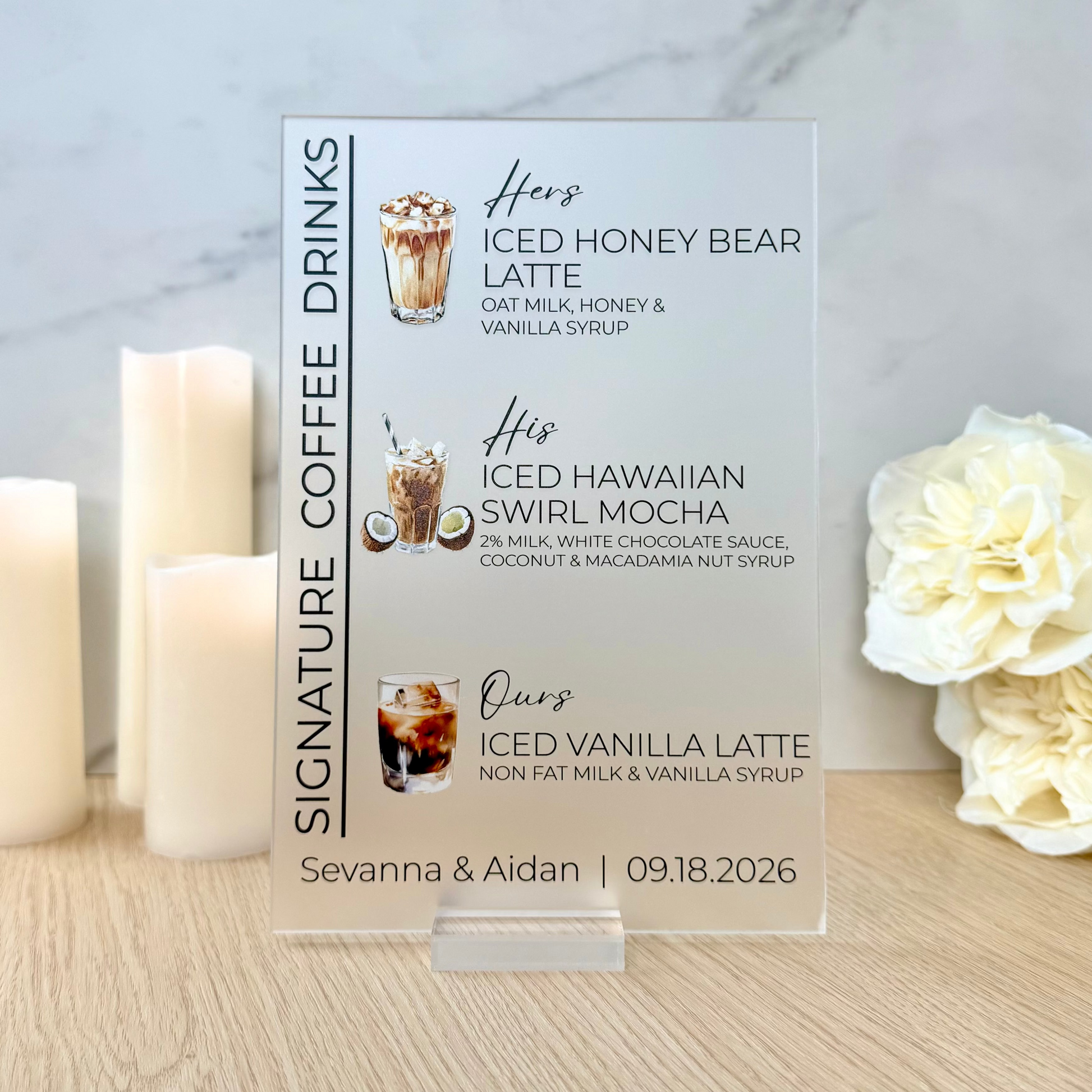 A clear acrylic custom coffee drink menu card standing on a wooden table, flanked by three white pillar candles on the left and white roses on the right, against a light marble wall.