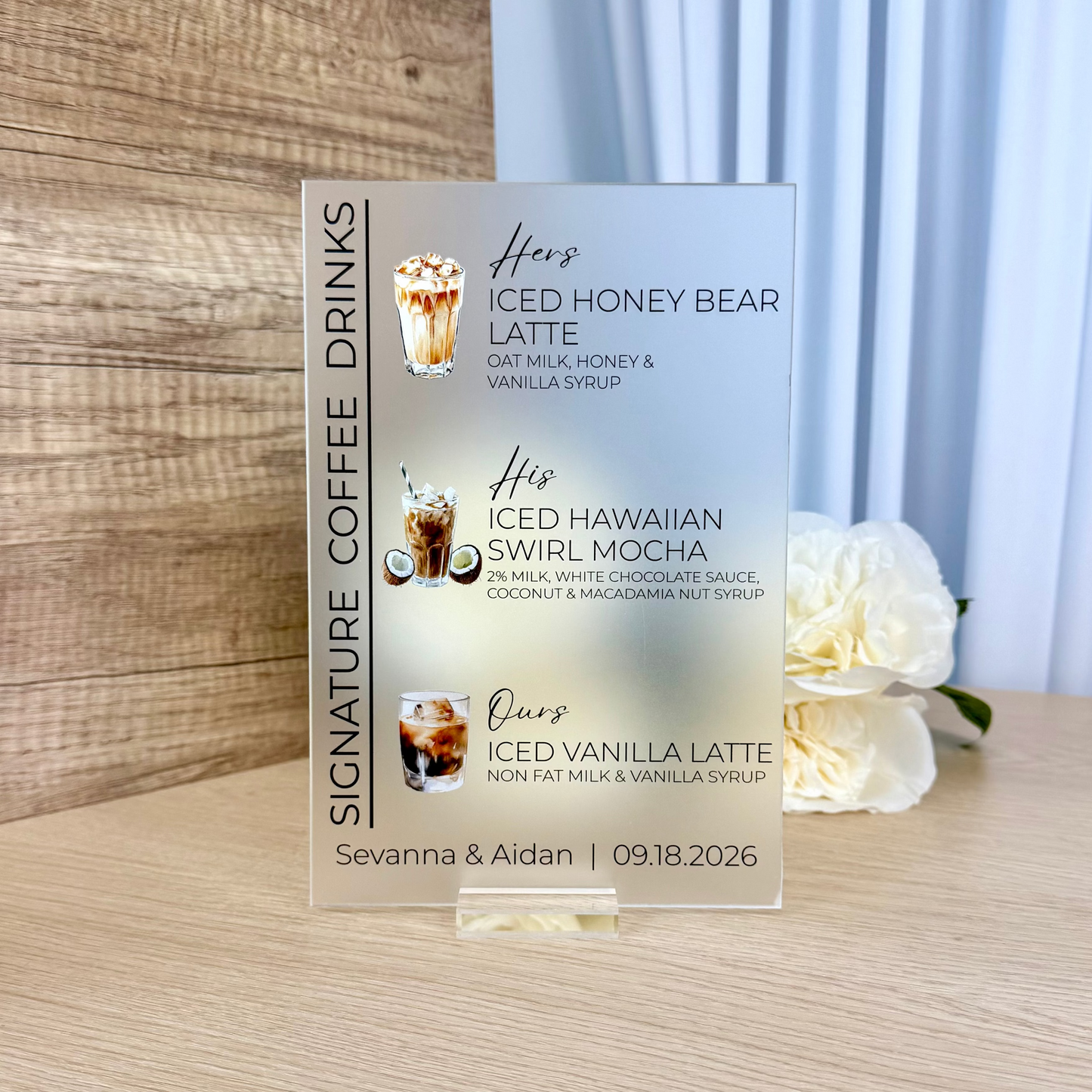 A custom acrylic sign displaying a personalized coffee menu with three iced drinks, standing on a light wood tabletop with a blurred background of wood paneling and white flowers.