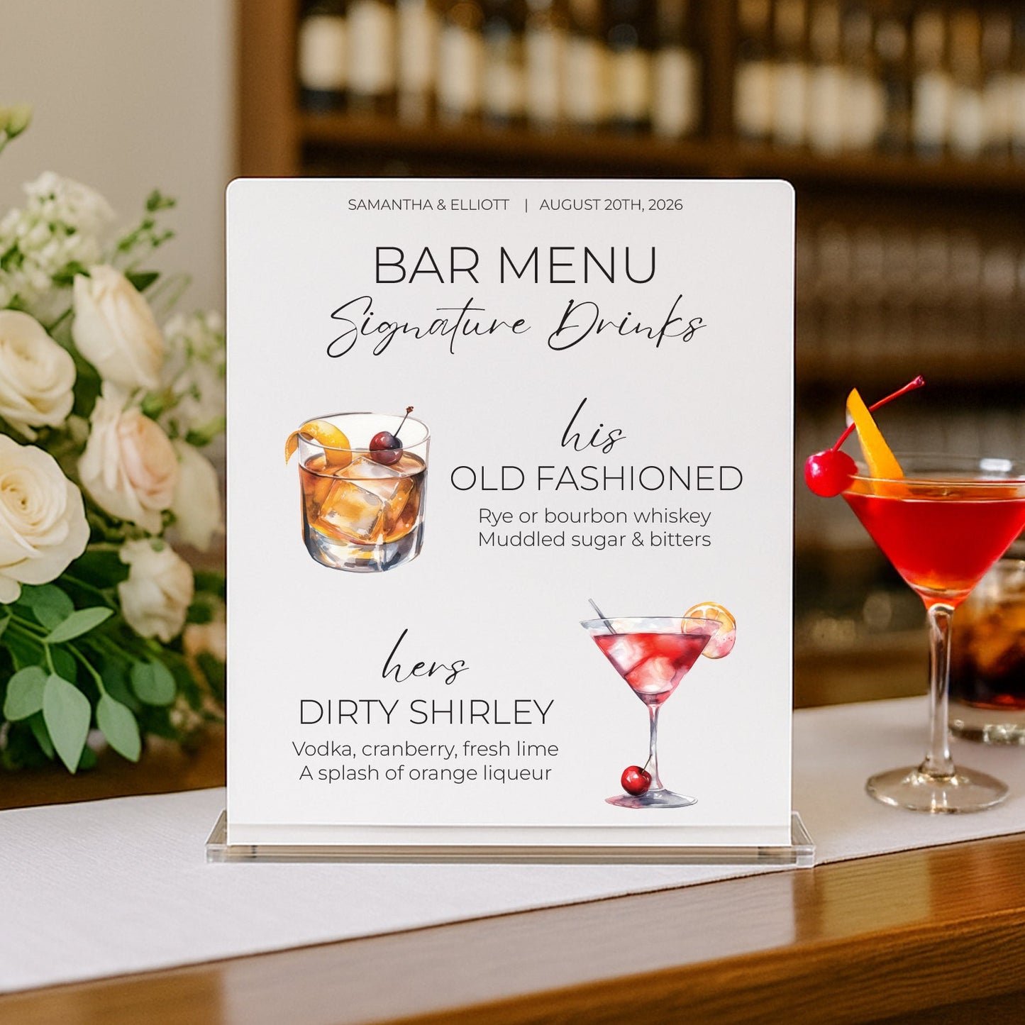 A clear acrylic bar menu sign displaying cocktail names and descriptions, styled on a wooden bar counter with a rose bouquet on the left and two cocktails on the right against a blurred bar background.